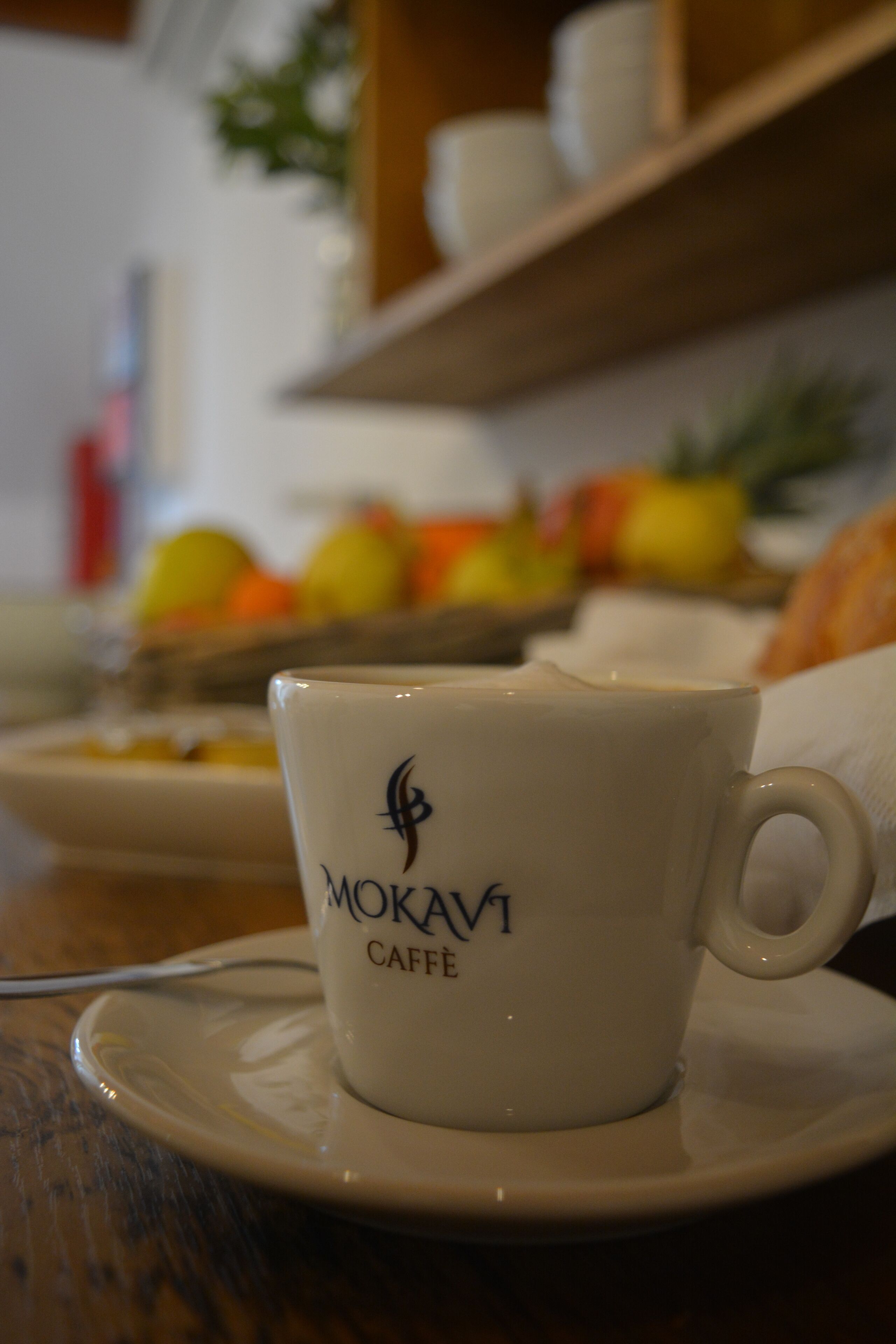 Una taza de café con un logo sobre un platito, colocada sobre una mesa con un fondo borroso de un cuenco de frutas y una estantería.