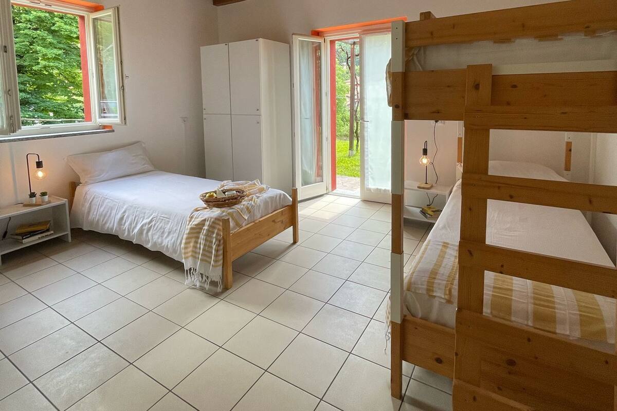 A bedroom with a wooden bunk bed, a single bed, a wardrobe, and a window with a view of greenery.