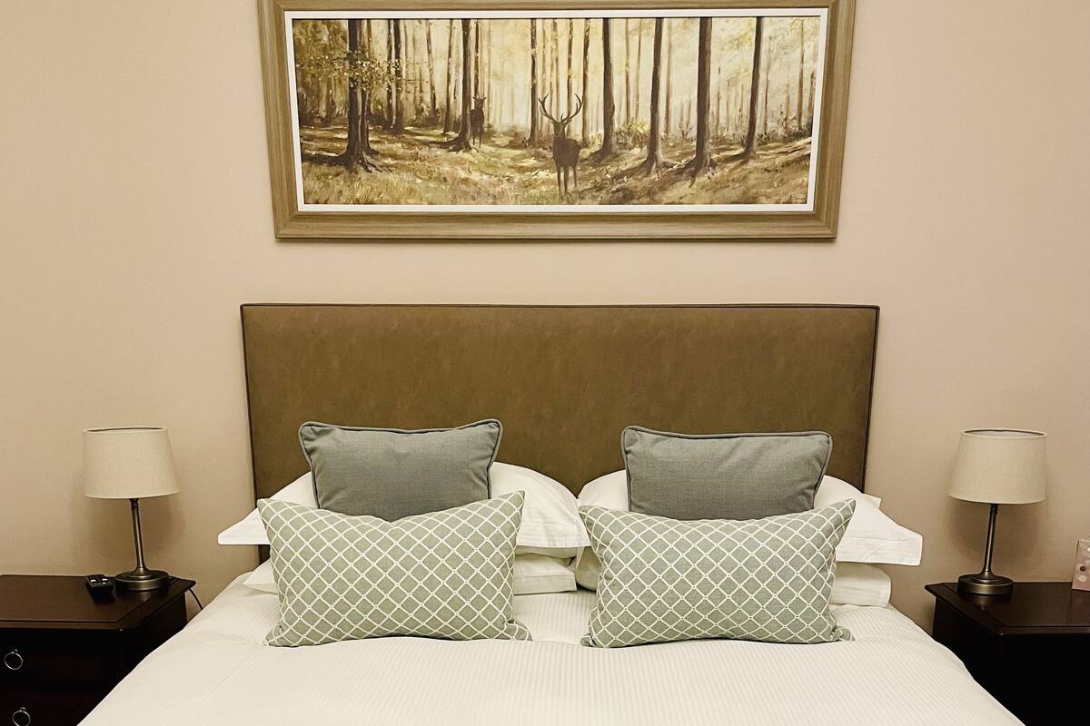 A neatly made bed with two pillows, a headboard, and a painting of a forest above it.