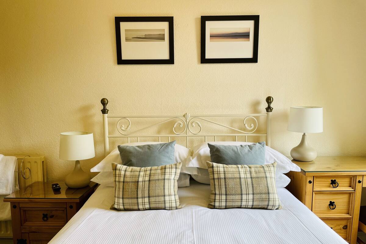 A neatly made bed with two pillows, a bedside table with a lamp, and a nightstand with a lamp. There are two framed pictures on the wall above the bed.