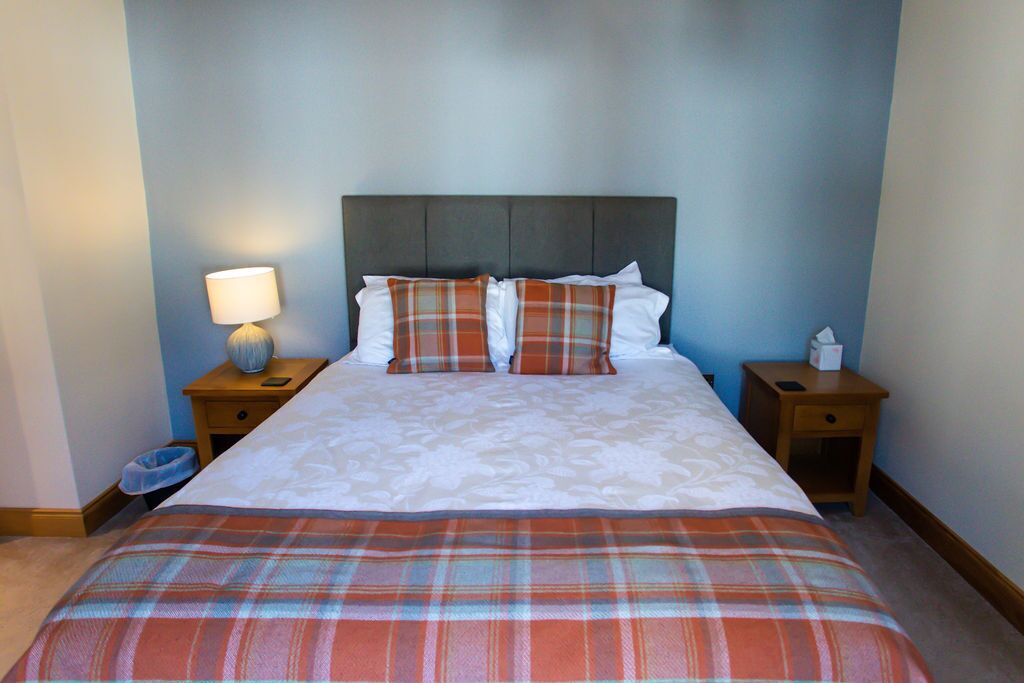 A neatly made bed with a plaid bedspread, two pillows, and a headboard. There are two bedside tables with lamps and a small trash bin.