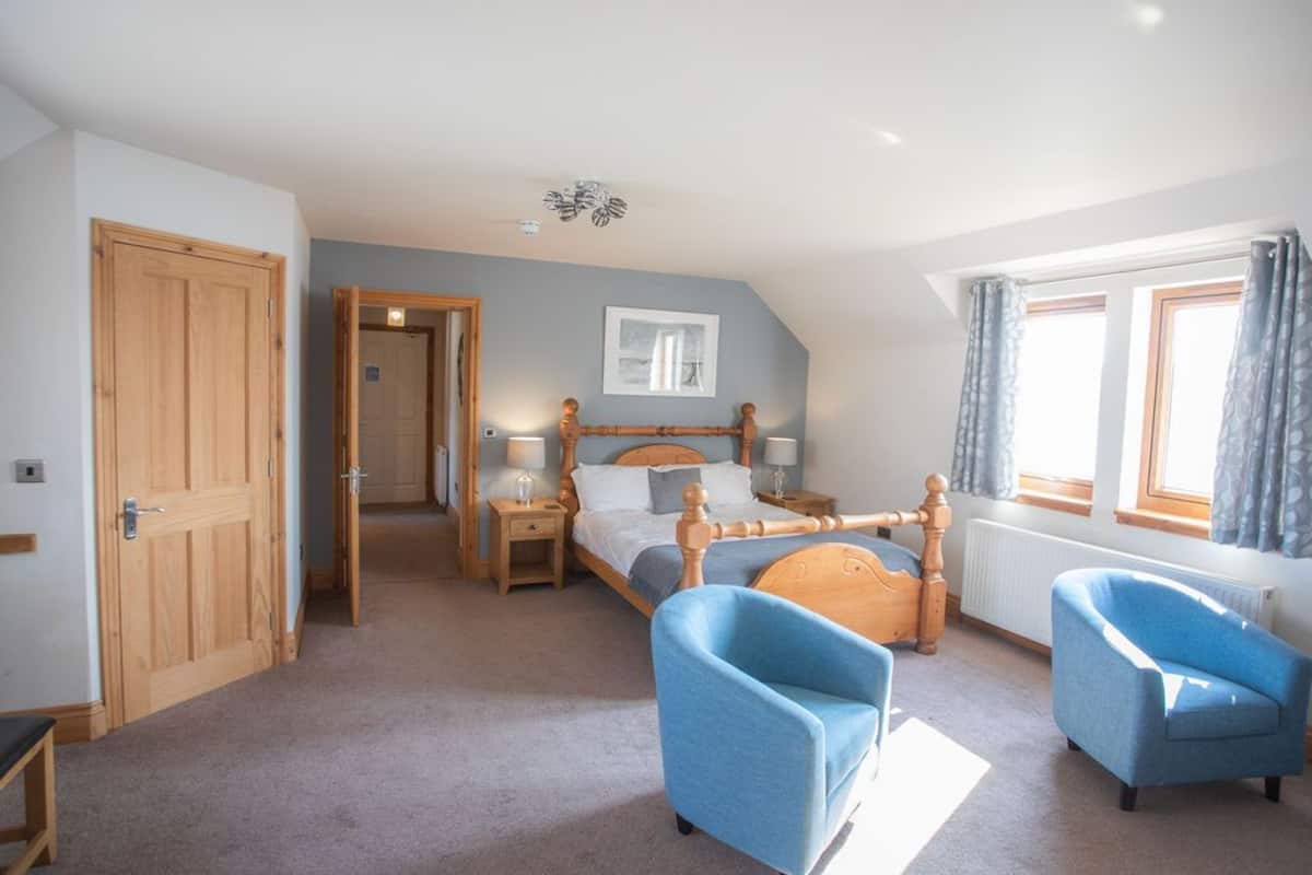 A bedroom with a wooden bed, two blue armchairs, a wooden wardrobe, and a window with curtains.