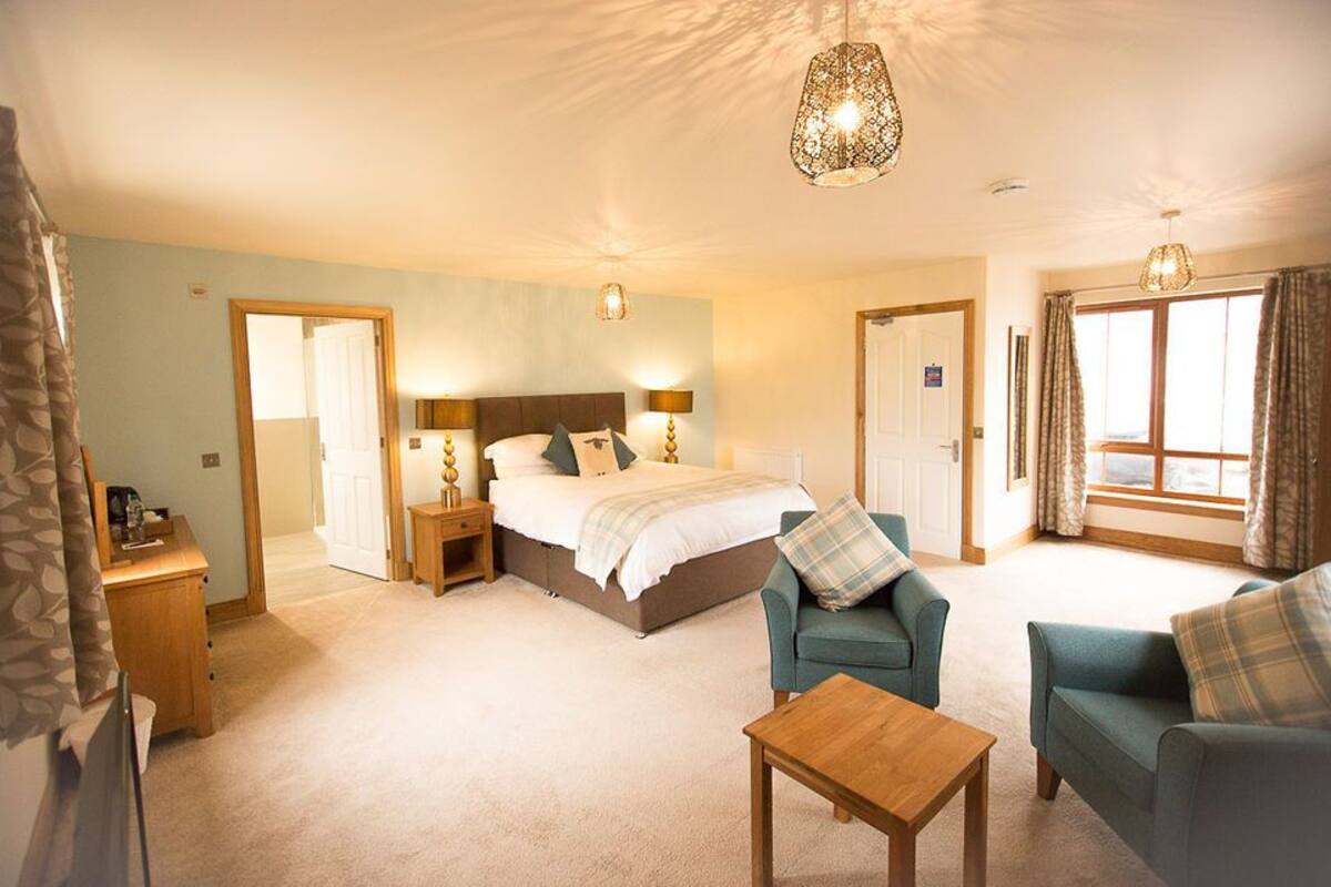 A hotel room with a bed, two bedside tables, a desk, two armchairs, a coffee table, and a large window with curtains.