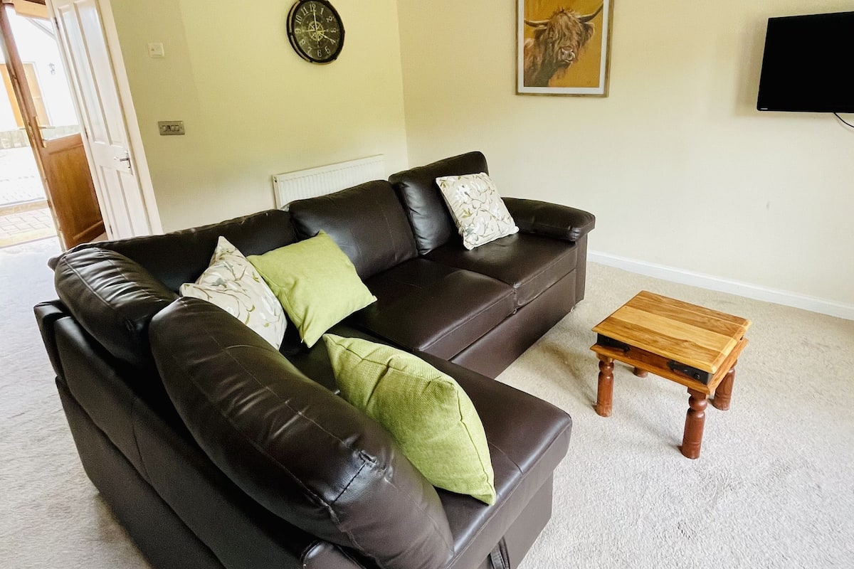 A living room with a dark brown leather sofa, two green pillows, a wooden coffee table, a wall clock, a framed picture, and a flat-screen TV.