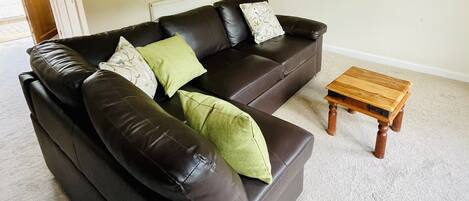 A living room with a dark brown leather sofa, two green pillows, a wooden coffee table, a wall clock, a framed picture, and a flat-screen TV.