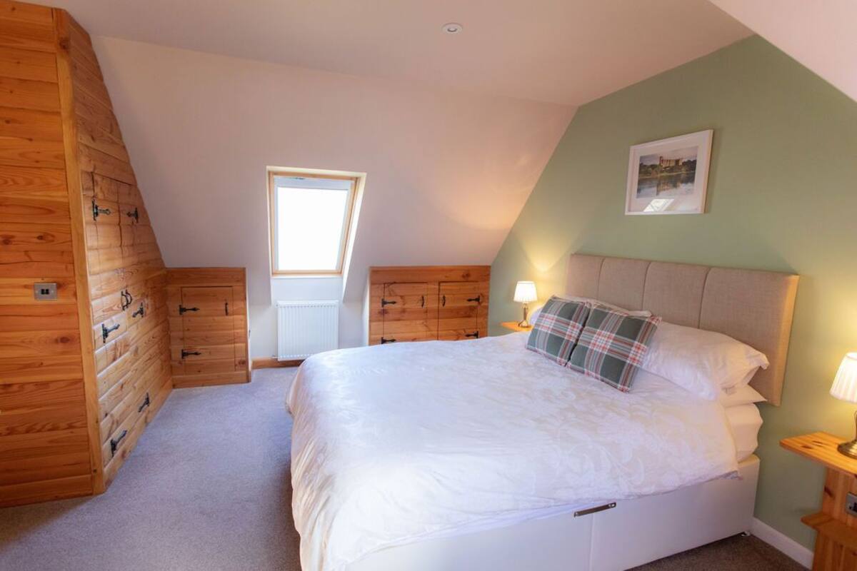 A bedroom with a sloped ceiling, wooden walls, a bed with white bedding, a plaid pillow, and a framed picture on the wall.