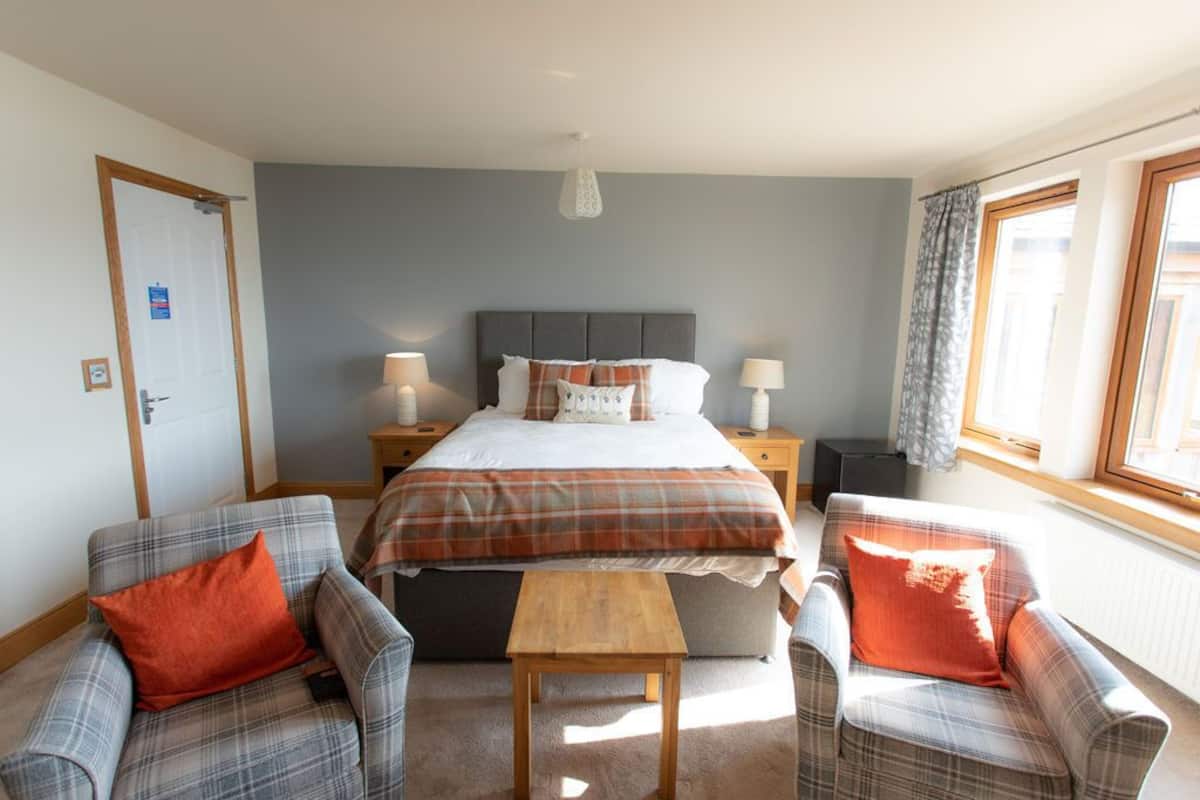 A bedroom with a bed, two nightstands, a wooden table, two plaid armchairs, and a window with curtains.
