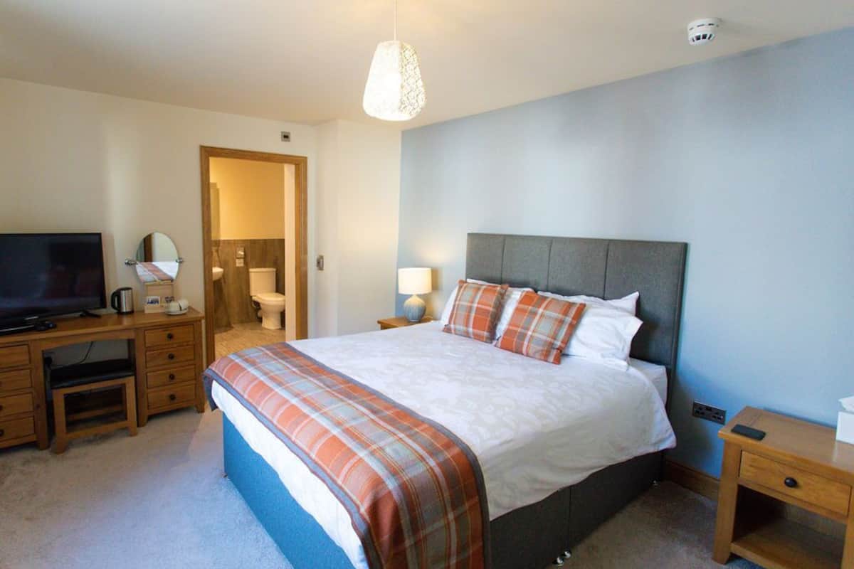 A bedroom with a bed, a desk with a television, a lamp, and a bedside table.