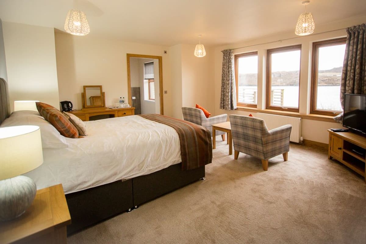 A bedroom with a bed, bedside table, lamp, chair, TV, and windows with curtains.