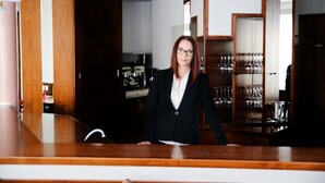 A reception area with a wooden counter, a woman in a black blazer, and a wine rack in the background. - Hotel Krone (Freilassing)