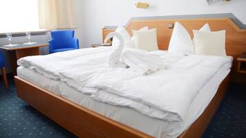 Ein ordentlich bezogenes Bett mit weißen Leinen und ein zu einem Schwan geformtes Handtuch, ein blauer Stuhl, ein Nachttisch mit einem Glas Wasser und ein Beistelltisch mit einer Lampe.