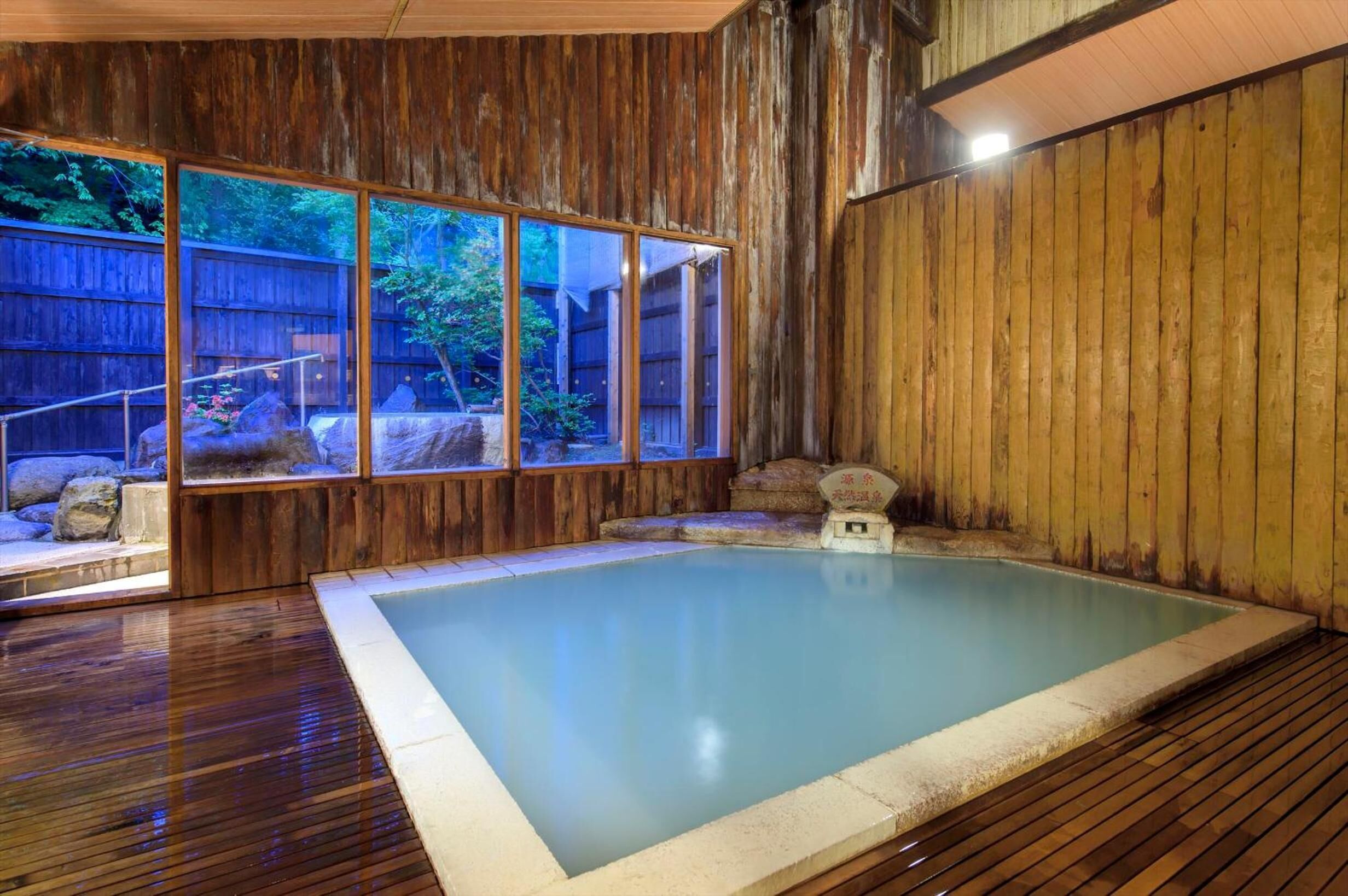 Interior de madera con un baño de aguas termales, paredes de madera y vistas a un jardín a través de amplios ventanales.