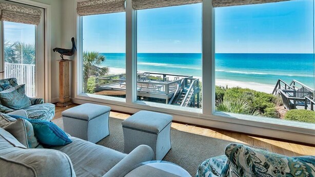 A room with a large window overlooking a beach, a balcony, and a view of the ocean.