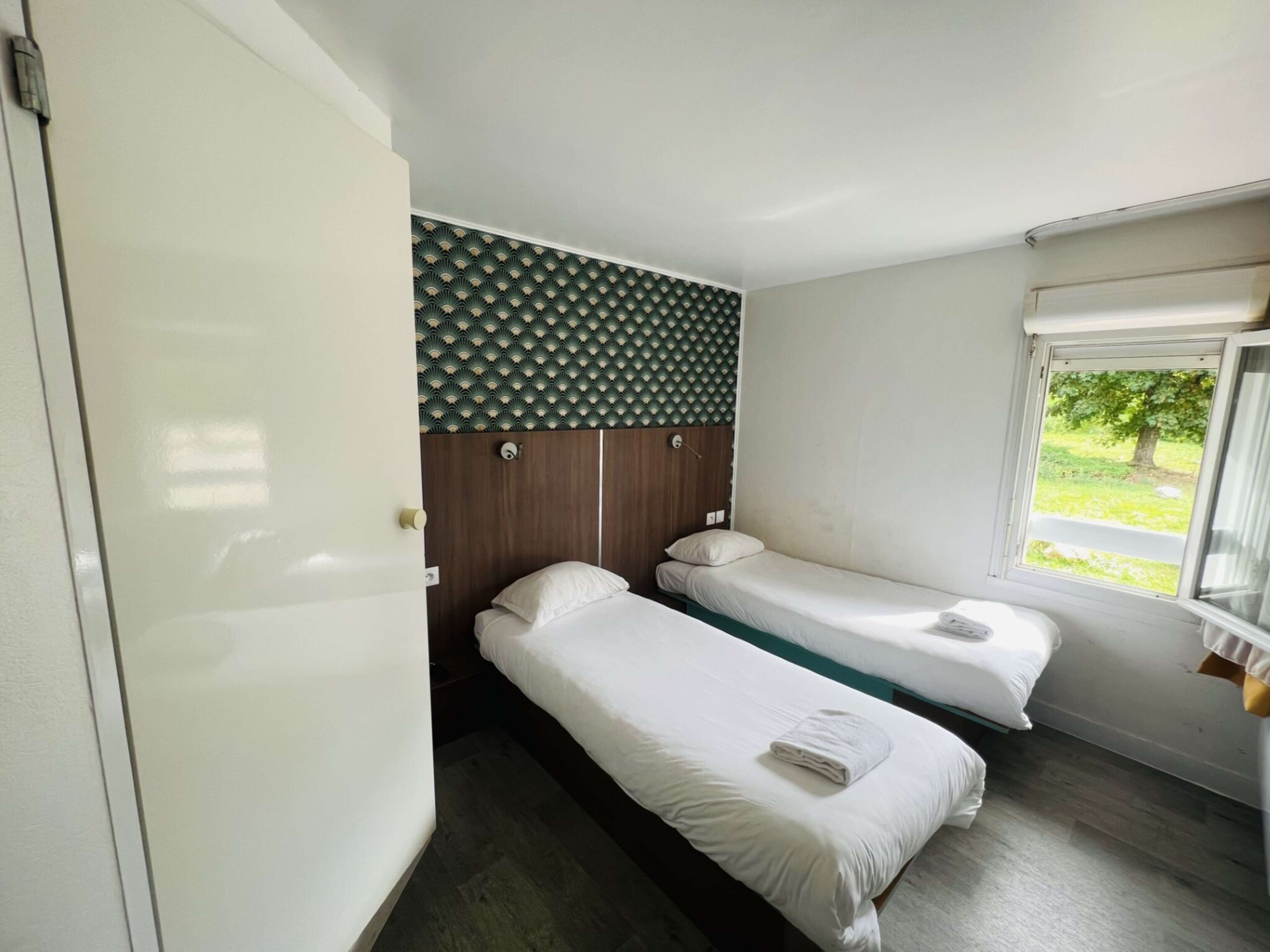 Habitación de hotel con dos camas individuales, cabecera de madera, ventana con vistas a vegetación y una puerta.
