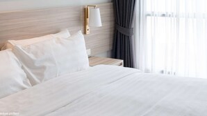 Un lit bien fait, avec du linge de lit blanc, une tête de lit en bois, une table de chevet avec une lampe et une fenêtre avec des rideaux.