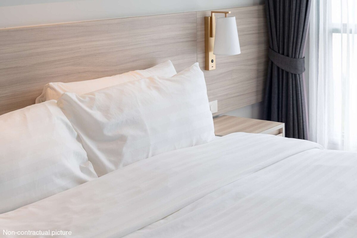 Una cama bien hecha con sábanas blancas, cabecera de madera, mesita de noche con lámpara y ventana con cortinas.