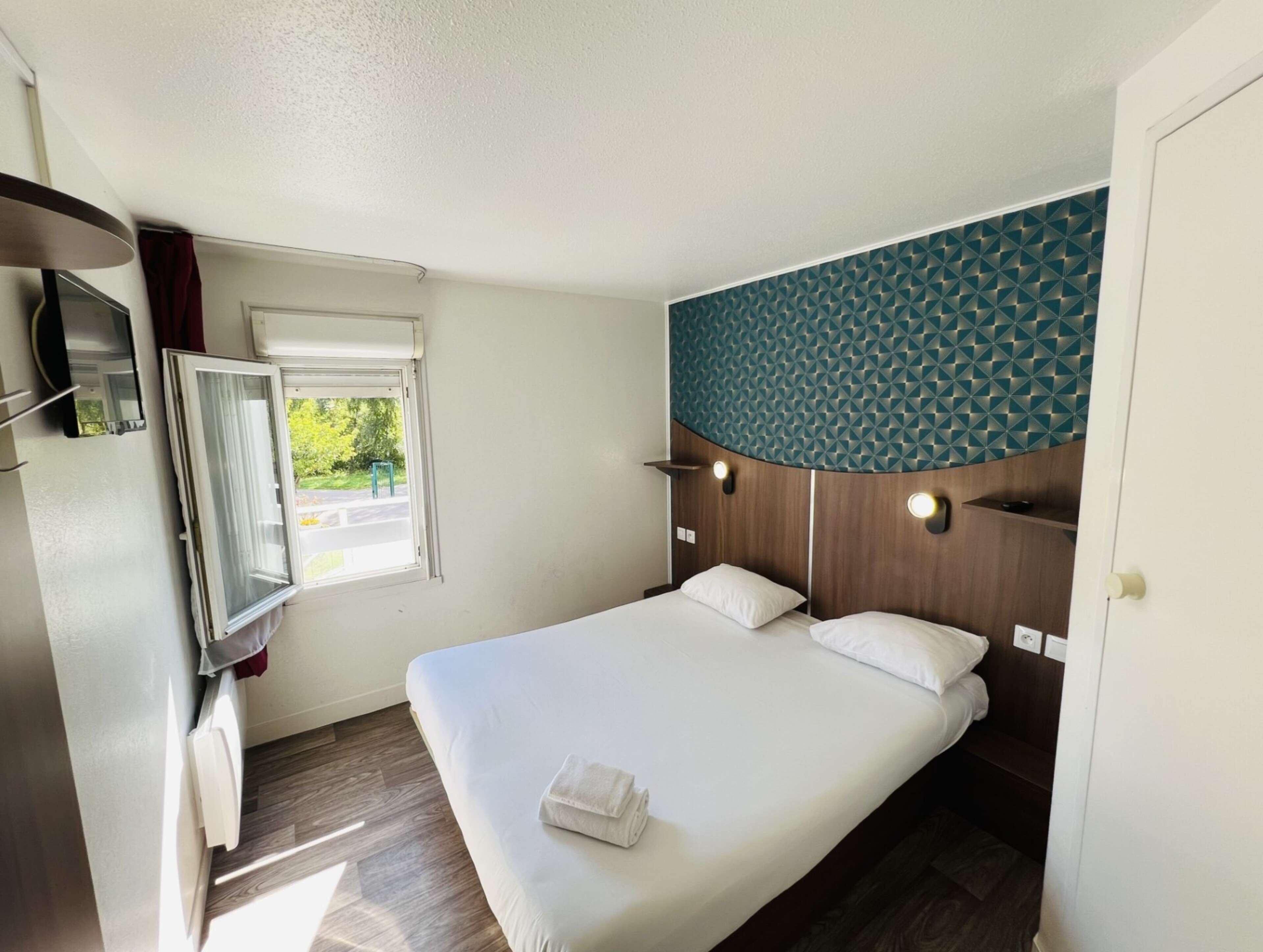 Habitación de hotel con cama, ventana con cortinas, televisión montada en la pared y cabecero de madera con diseño geométrico.