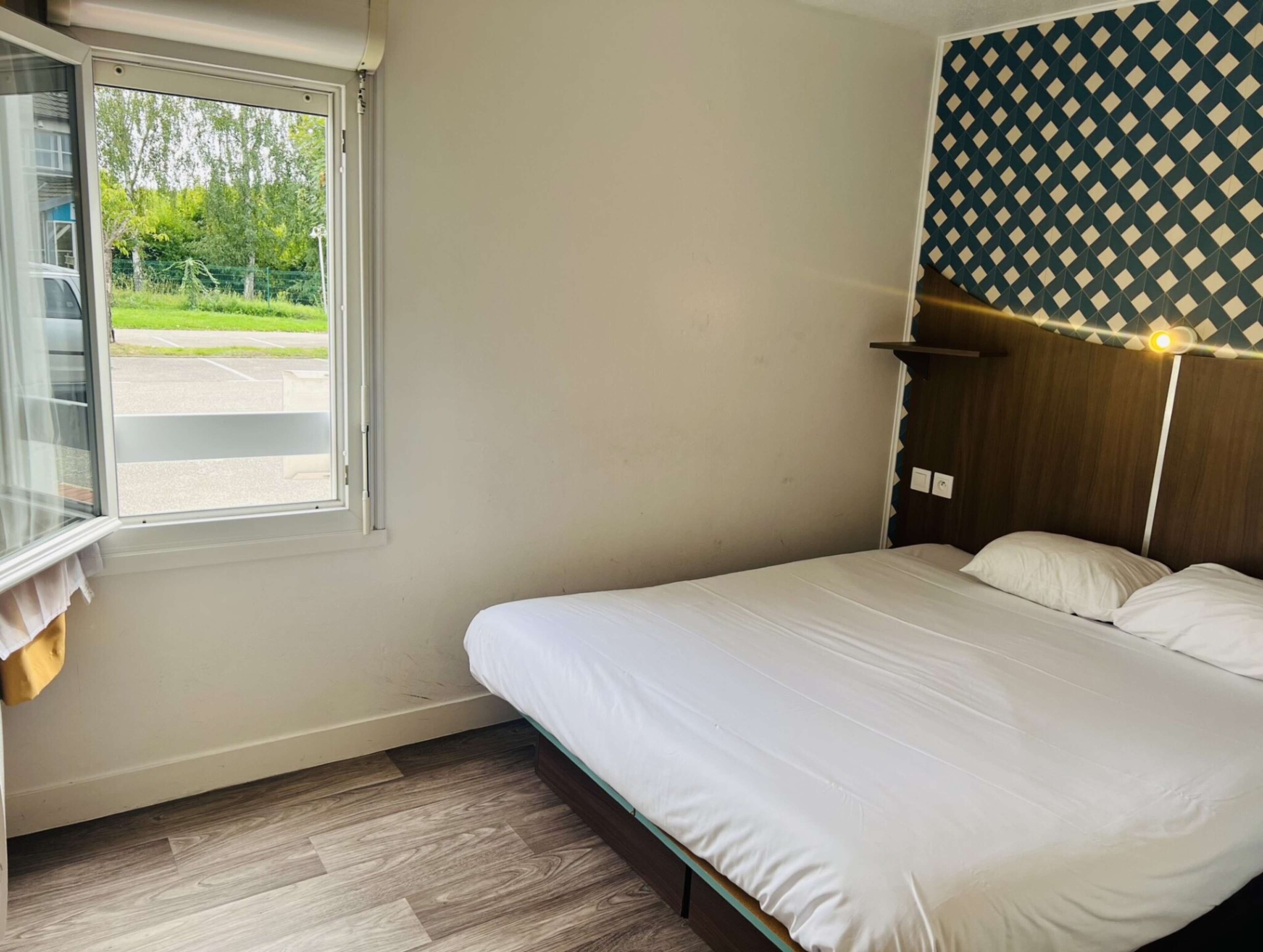 Un lit simple avec une tête de lit en bois, une fenêtre donnant sur un parking et sur de la verdure, et un mur décoré d’un motif.