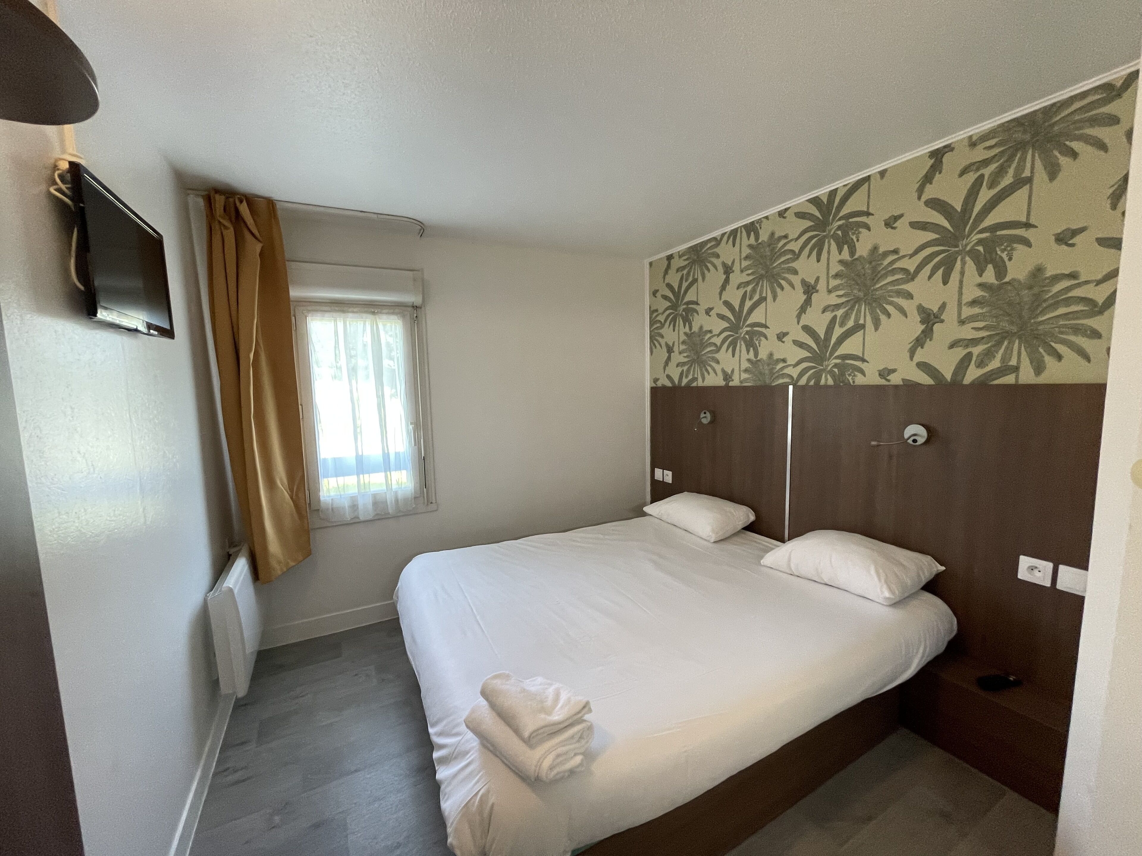 Un lit simple avec du linge de lit blanc, un téléviseur fixé au mur, une fenêtre avec des rideaux et un papier peint à motifs floraux.
