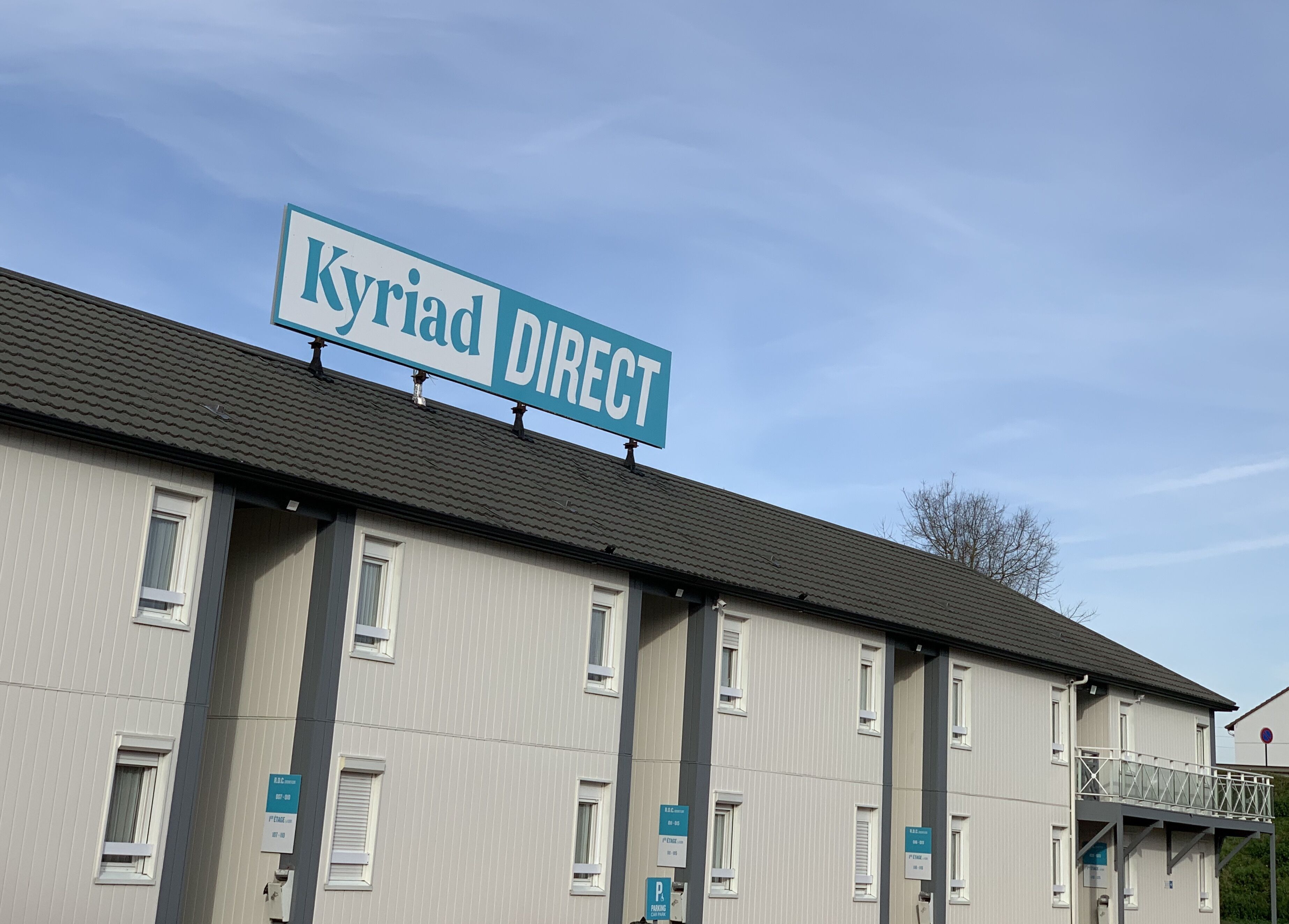 Ein Gebäude mit einer Beschilderung mit dem Schriftzug "Kyriad DIRECT“ und zahlreichen Fenstern mit Schildern, die Zimmernummern und Annehmlichkeiten angeben.