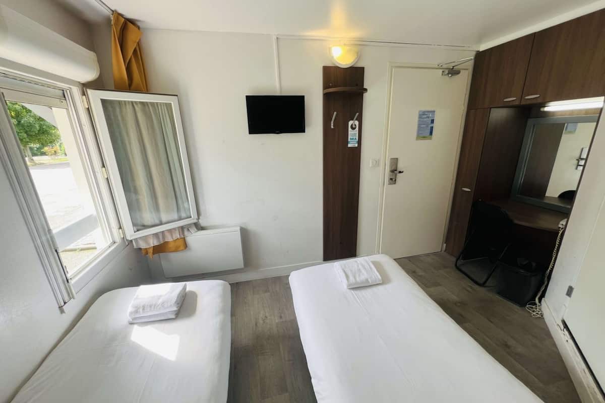 Ein kleines, sauberes Zimmer mit zwei Einzelbetten, einem kleinen Fenster mit Vorhängen, einem Spiegel und einem Holzschrank.