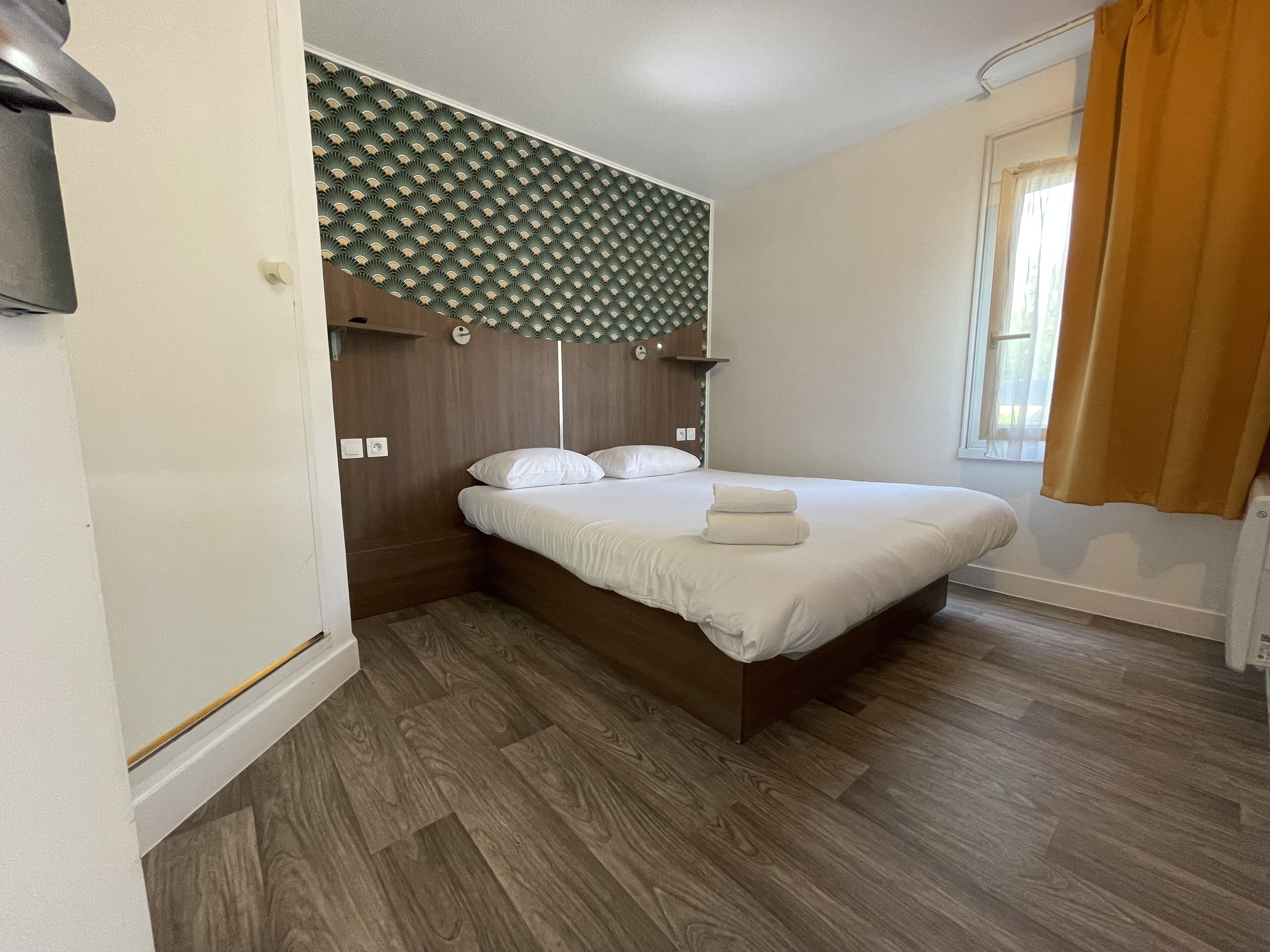 A neatly made bed with white linens, a wooden headboard, and a wall-mounted light fixture above it.