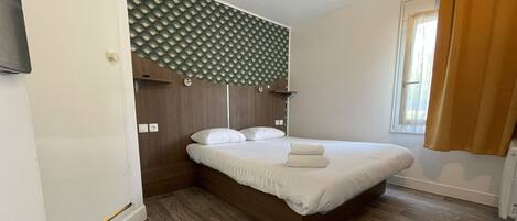 A neatly made bed with white linens, a wooden headboard, and a wall-mounted light fixture above it.