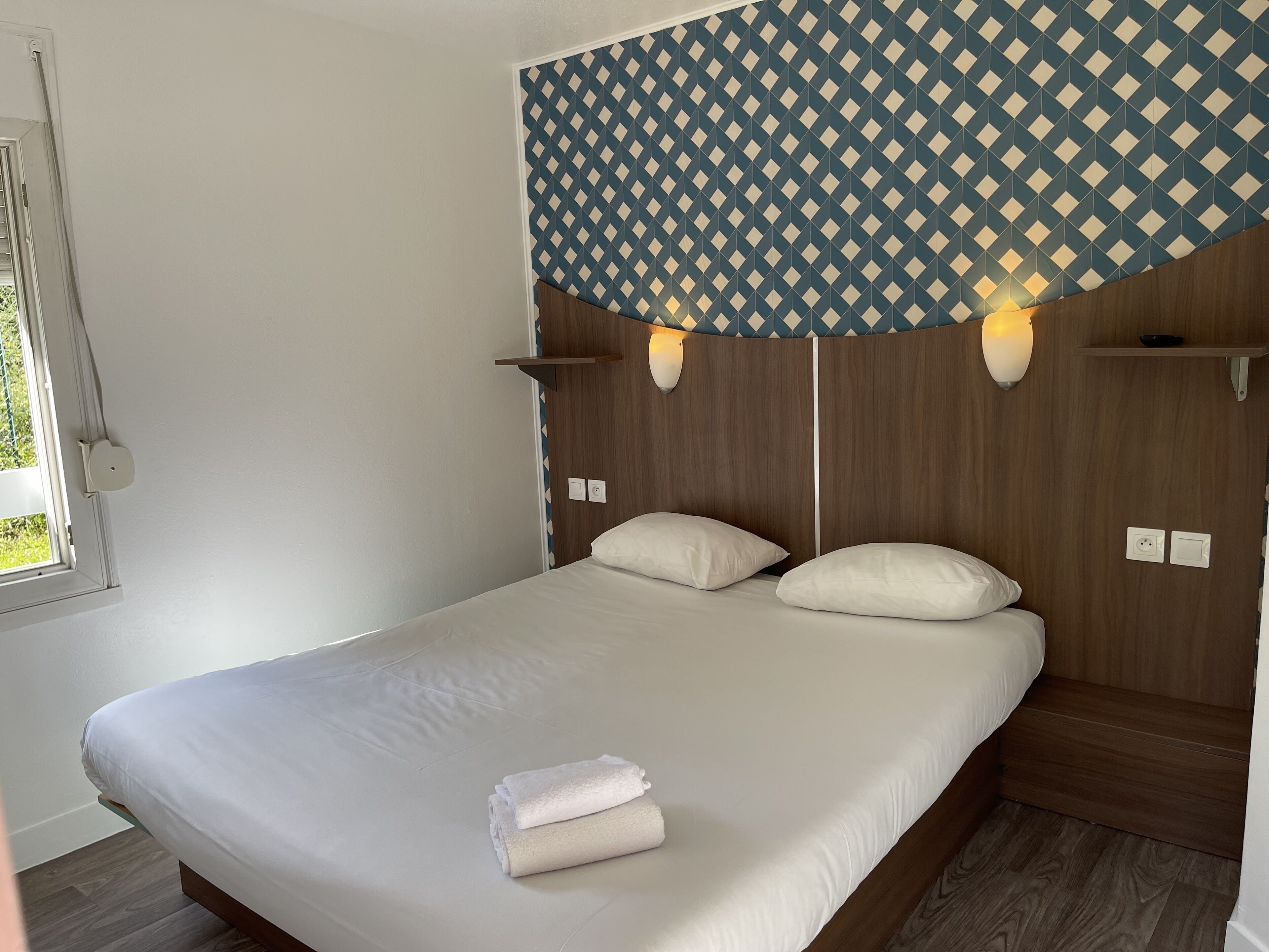 Una cama bien hecha con sábanas blancas y una toalla doblada, cabecera de madera y cortinas con estampado geométrico, además de lámparas de noche.