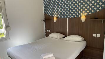 Una cama bien hecha con sábanas blancas y una toalla doblada, cabecera de madera y cortinas con estampado geométrico, además de lámparas de noche.
