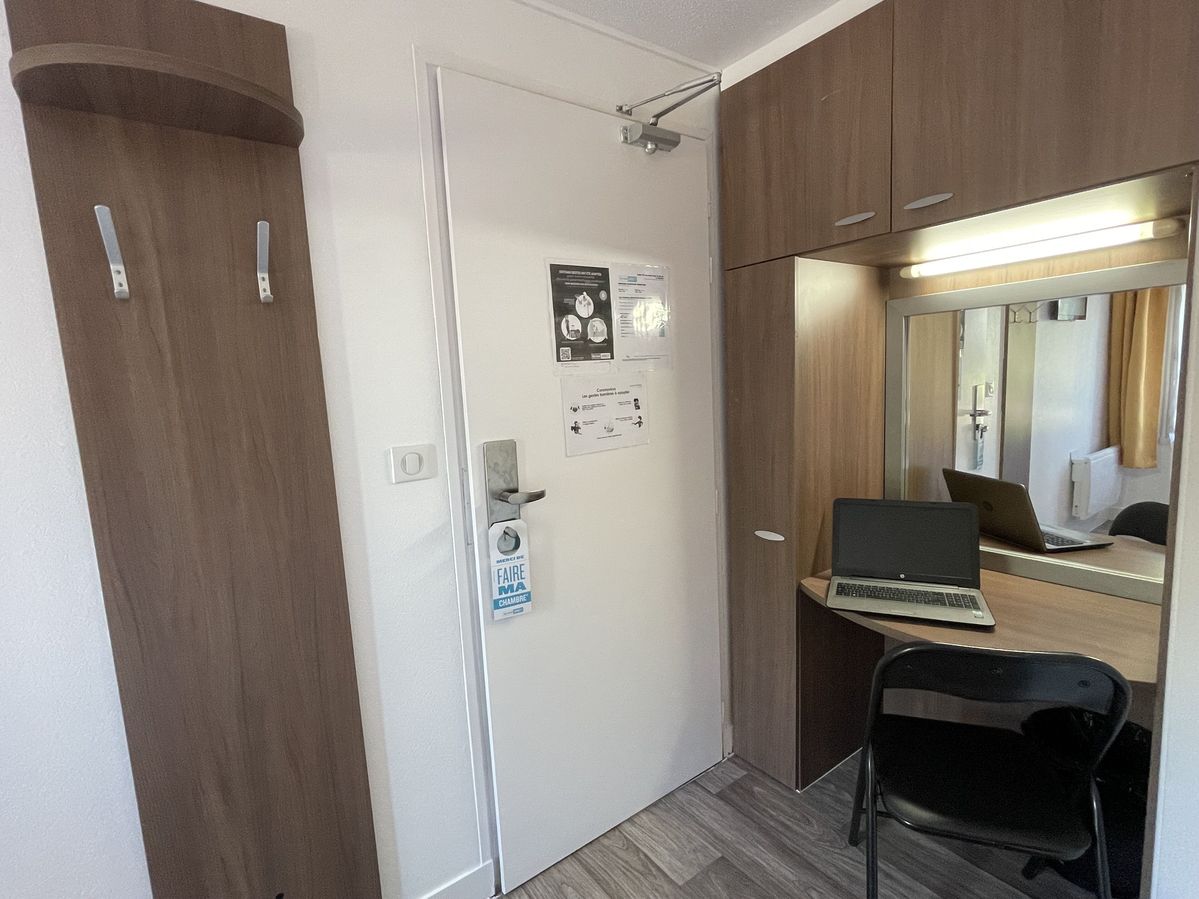 A compact room with a wooden wardrobe, a desk with a laptop, a chair, and a mirror.