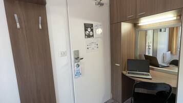 A compact room with a wooden wardrobe, a desk with a laptop, a chair, and a mirror.