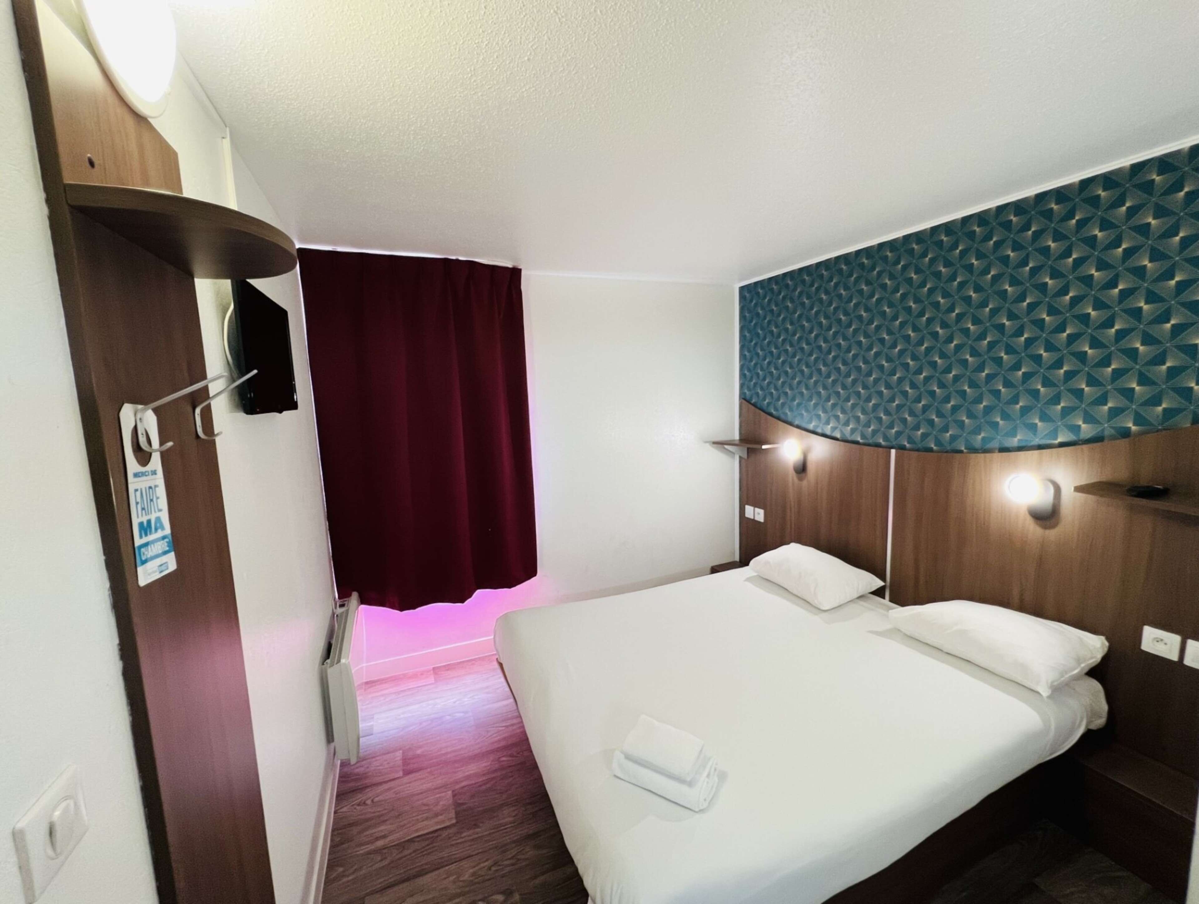 A hotel room with a bed, a wooden headboard, a red curtain, and a wall with a geometric pattern.