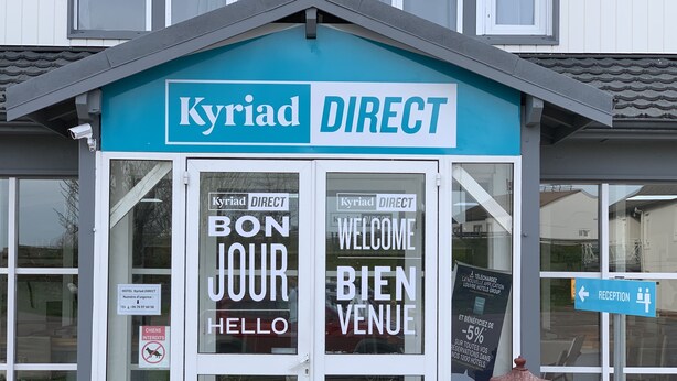 L’entrée d’un hôtel avec une enseigne « Kyriad DIRECT », un panneau d’accueil et une vitrine avec l’inscription « BON JOUR HELLO ».