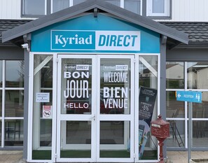 L’entrée d’un hôtel avec une enseigne « Kyriad DIRECT », un panneau d’accueil et une vitrine avec l’inscription « BON JOUR HELLO ».