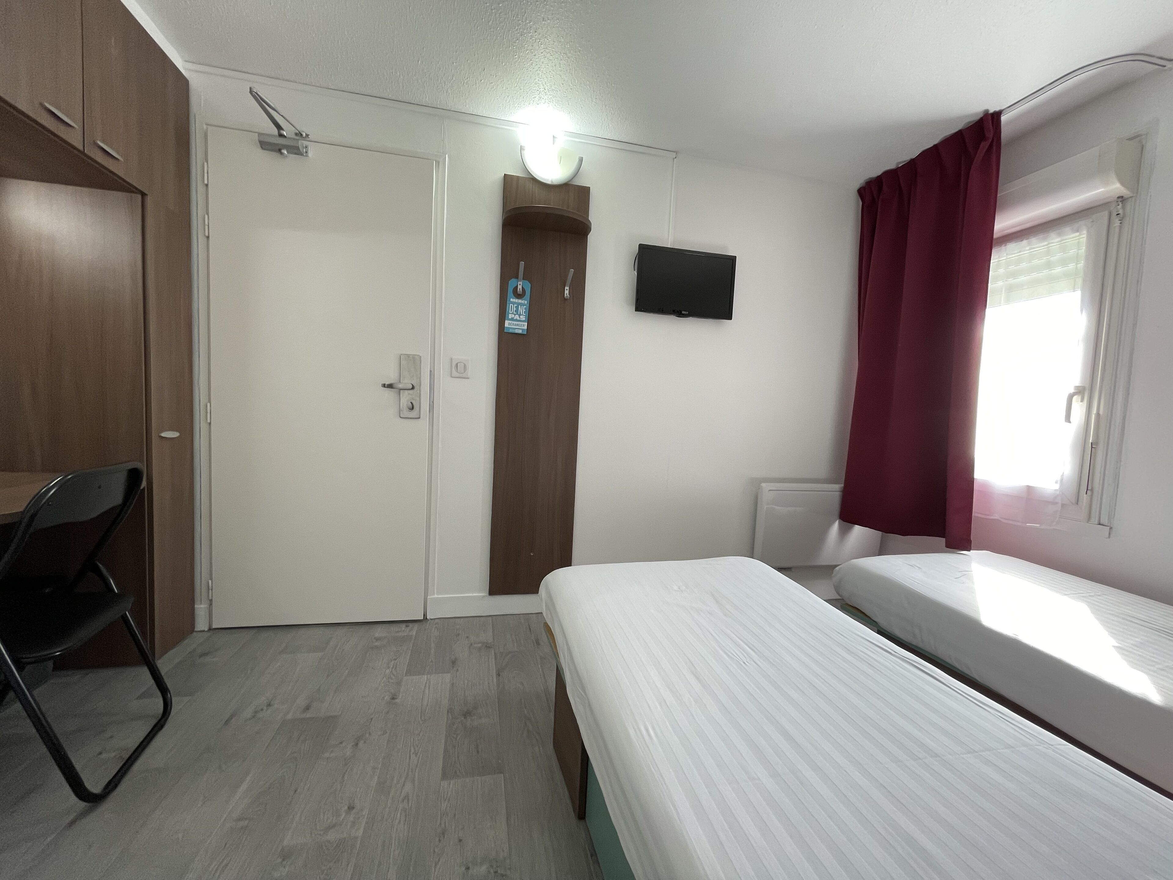 Un dormitorio con cama, escritorio y silla. Se ve una puerta con un cartel, un televisor en la pared y una ventana con cortinas.