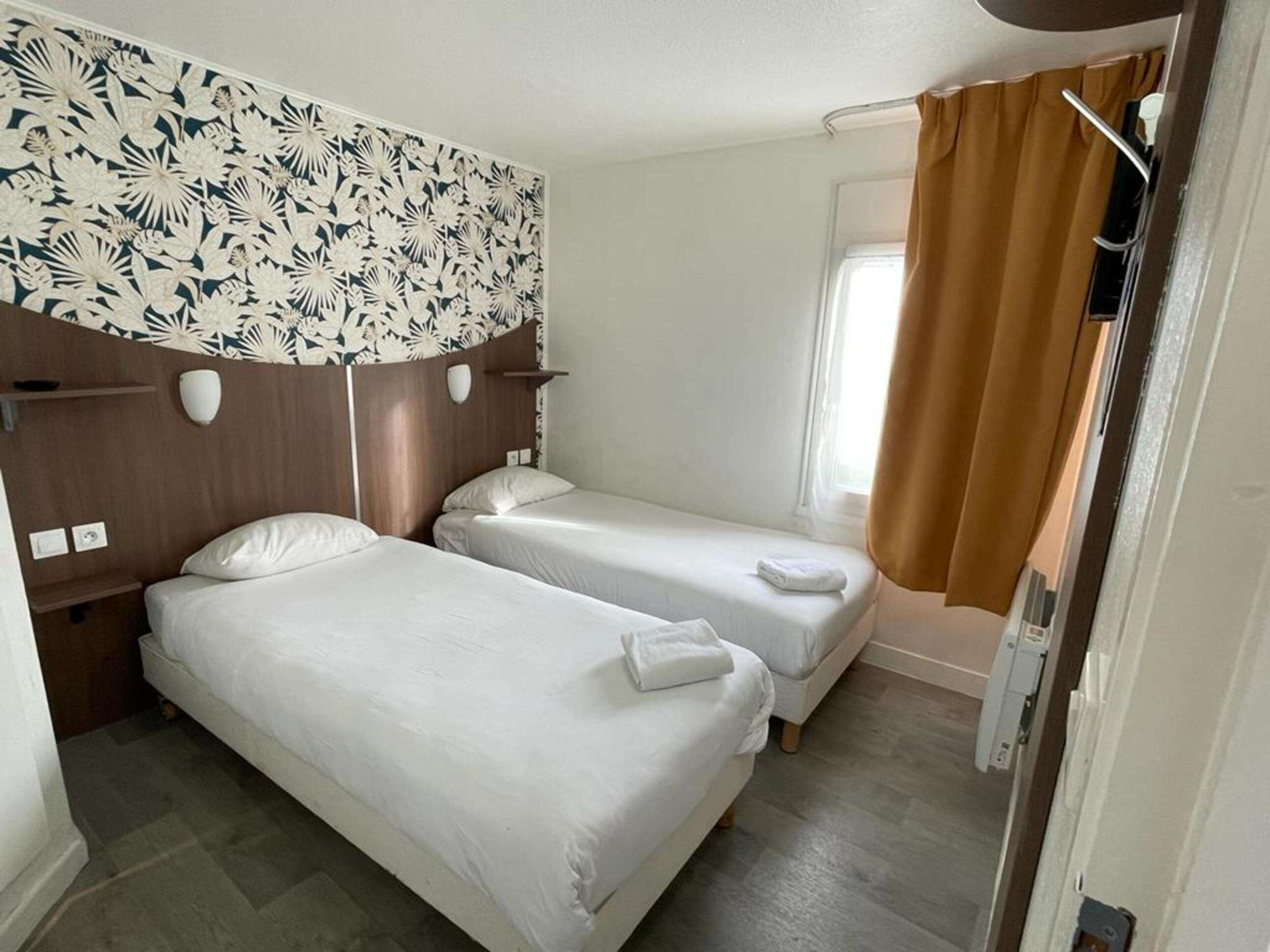 A hotel room with two single beds, a wooden headboard with a floral pattern, a window with brown curtains, and a wall-mounted light fixture.