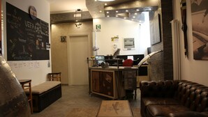 A cozy lounge area with a leather sofa, a wooden bench, and a small table. A chalkboard with various writings and a clock is on the wall. A kitchenette with a counter and cabinets is visible in the background. - Atmos Luxe Navigli Hostel & Rooms (Milan)