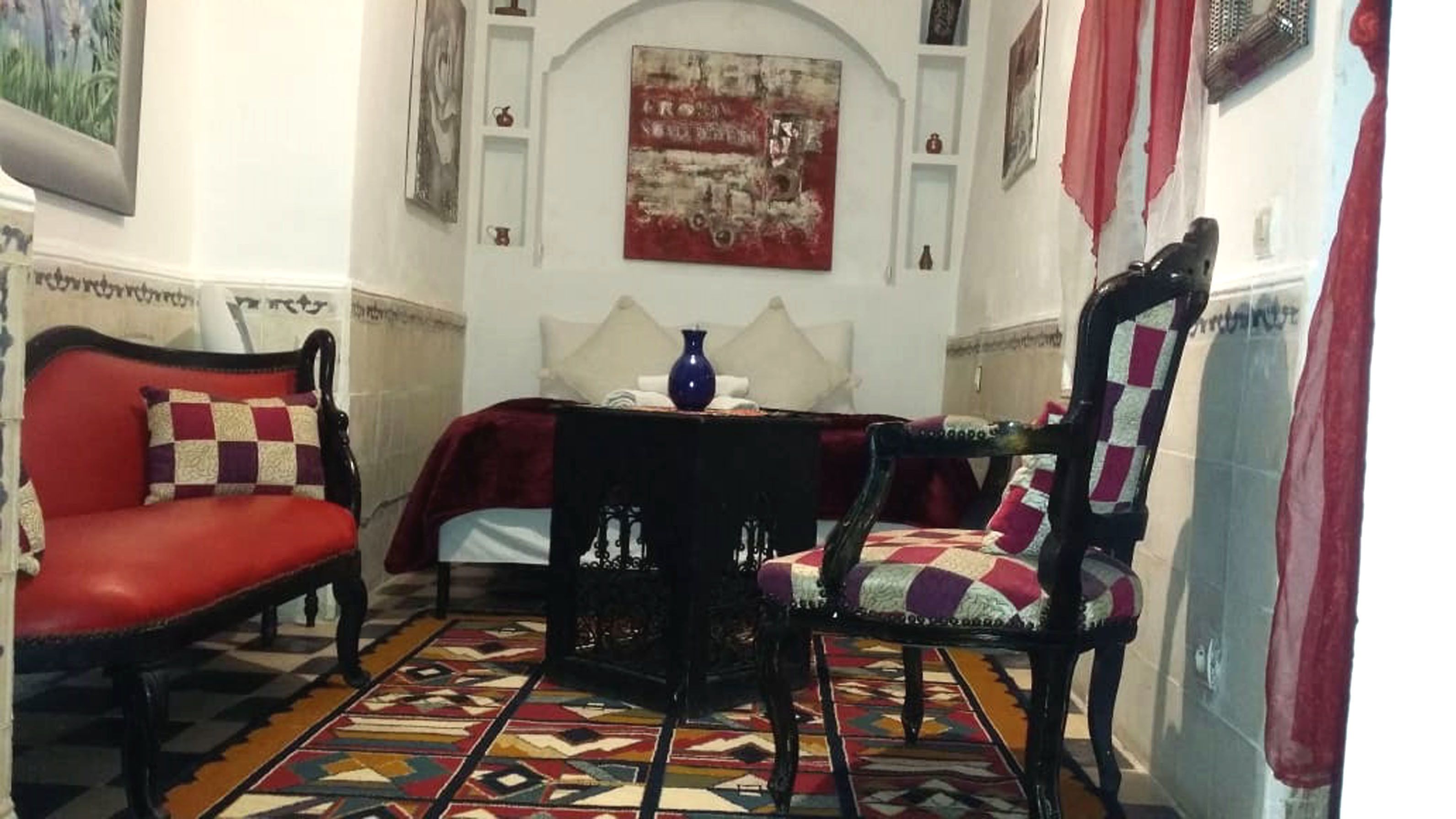 A room with a red sofa, a patterned armchair, a small table with a vase, and a colorful rug.