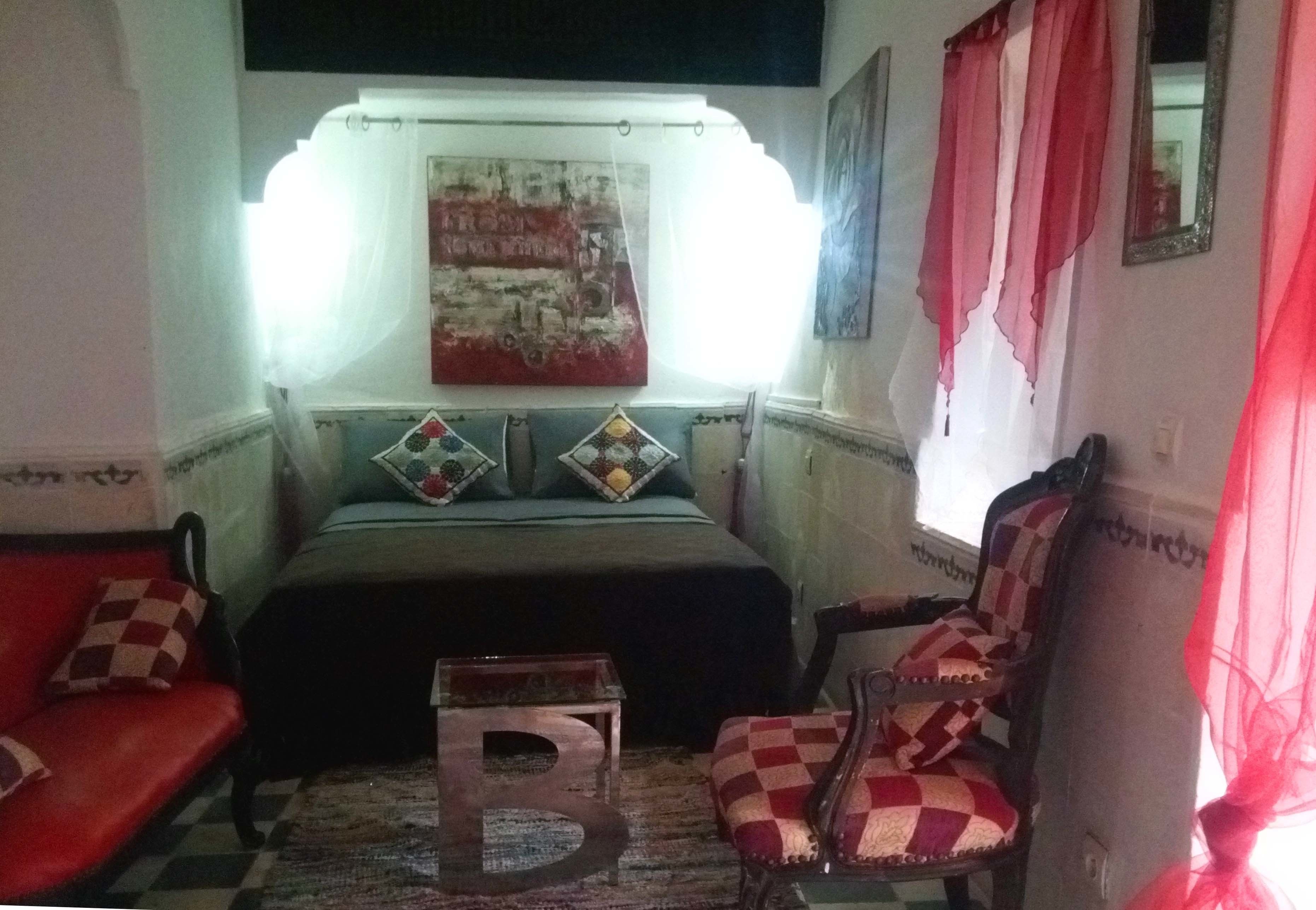 A room with a bed, a red sofa, a checkered armchair, a small wooden table, and a large abstract painting on the wall.
