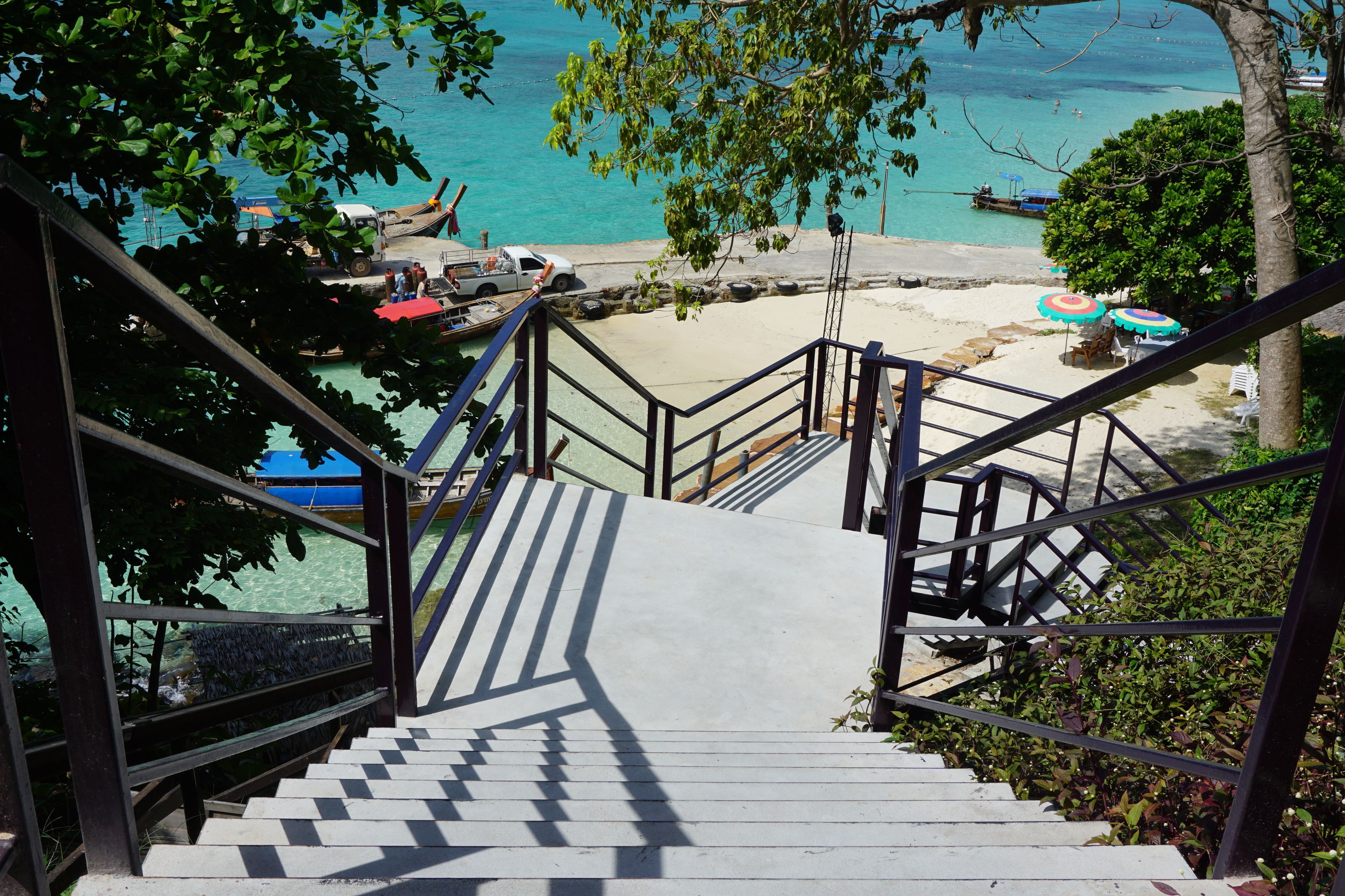 Eine Treppe führt zu einem Strandbereich mit klarem türkisblauem Wasser, Booten und Sonnenschirmen.