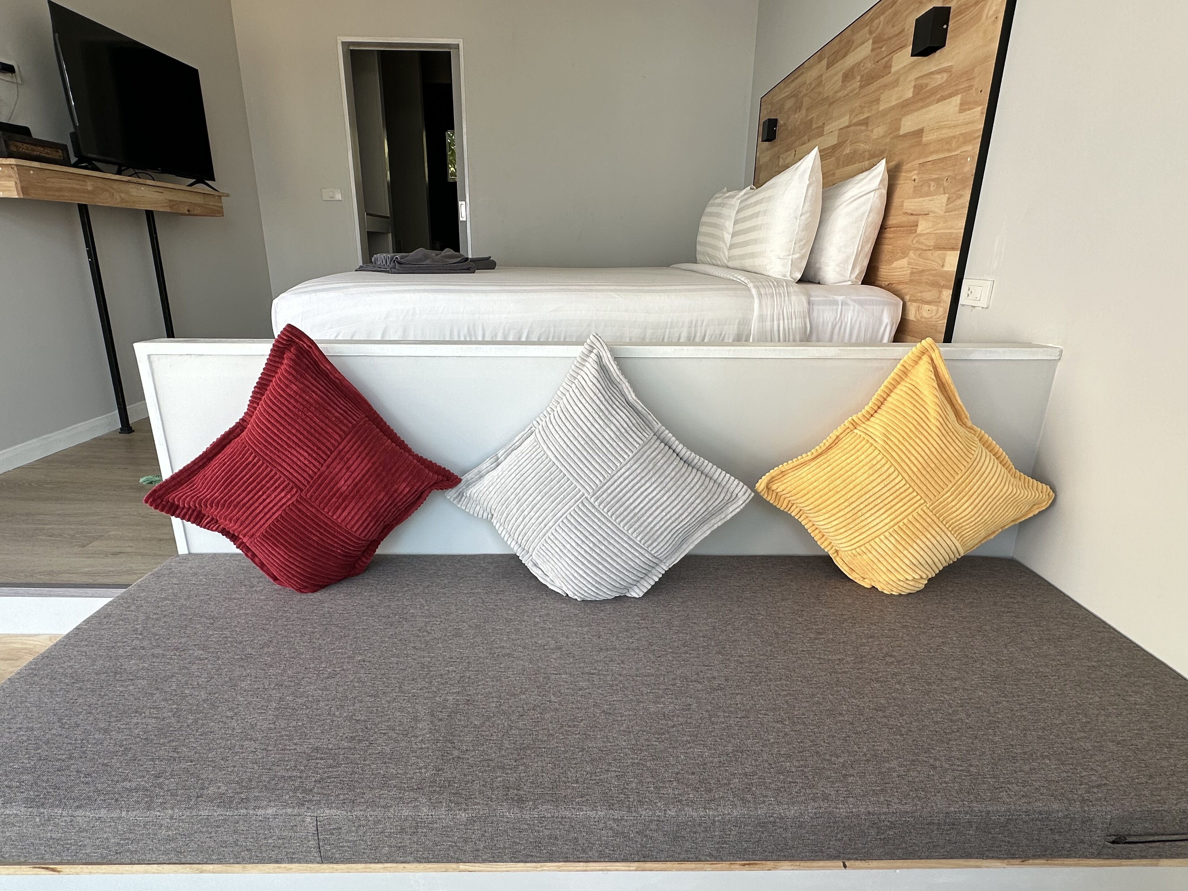 Une chambre à coucher moderne avec un lit, un téléviseur posé sur un meuble et trois coussins sur un pouf gris.