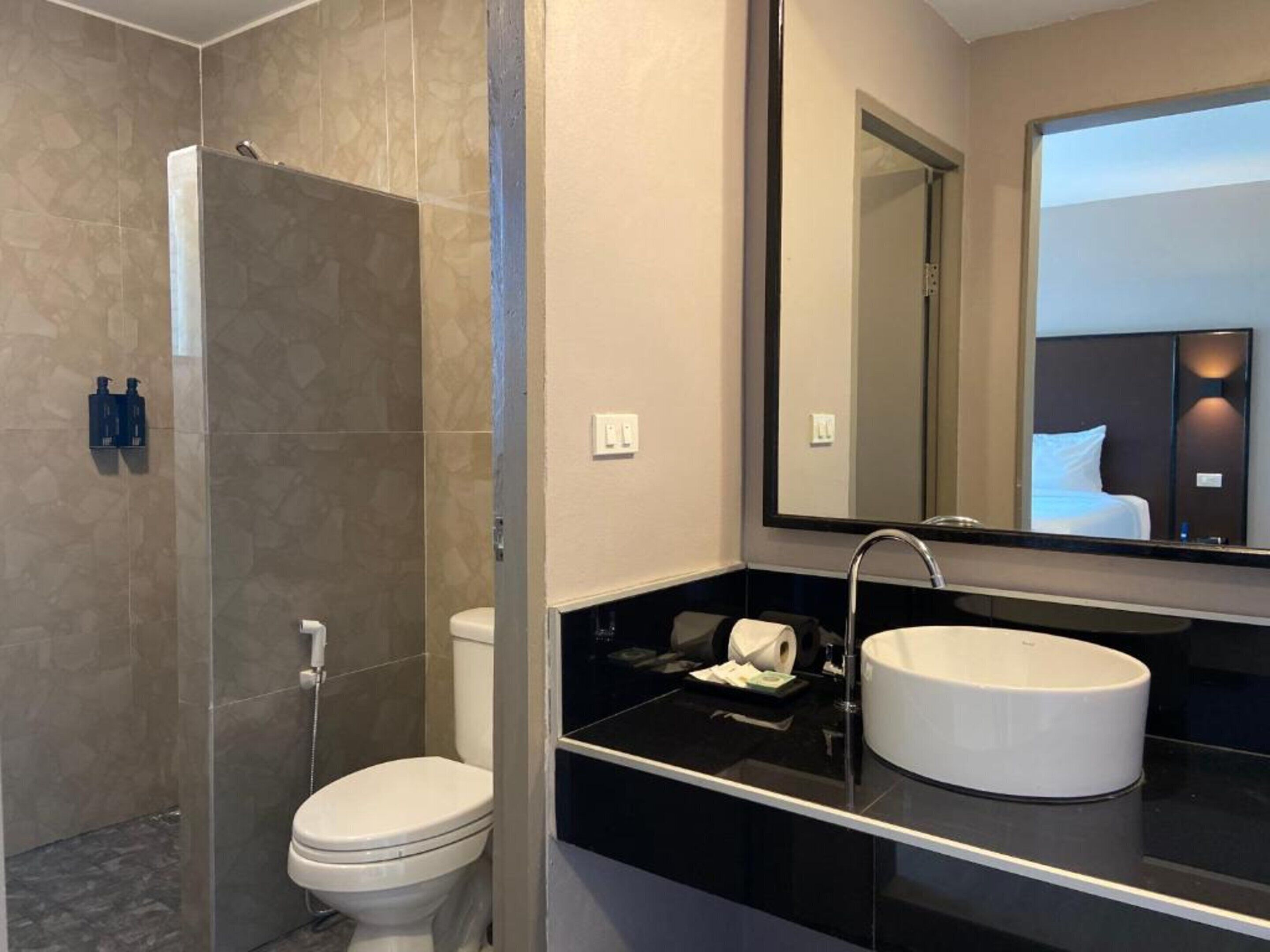 Un baño moderno con ducha envuelta en vidrio, inodoro blanco, encimera negra y un espejo grande que refleja una cama.
