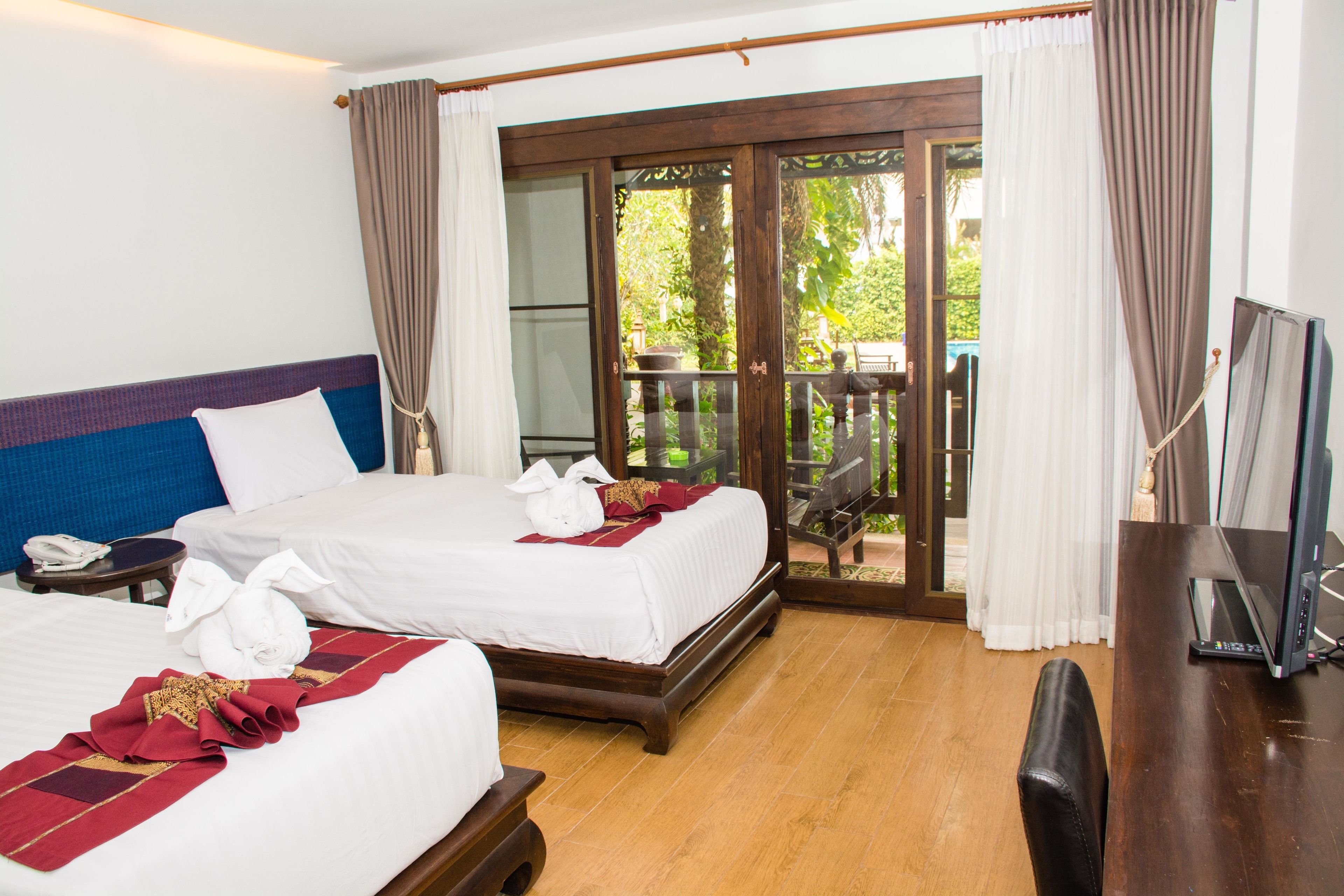 A hotel room with two beds, a desk, a TV, and a balcony with a view of greenery.