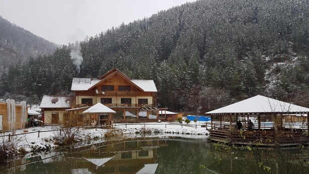 Un chalet en bois, avec un grand étang devant, entouré d’arbres et de collines couverts de neige.