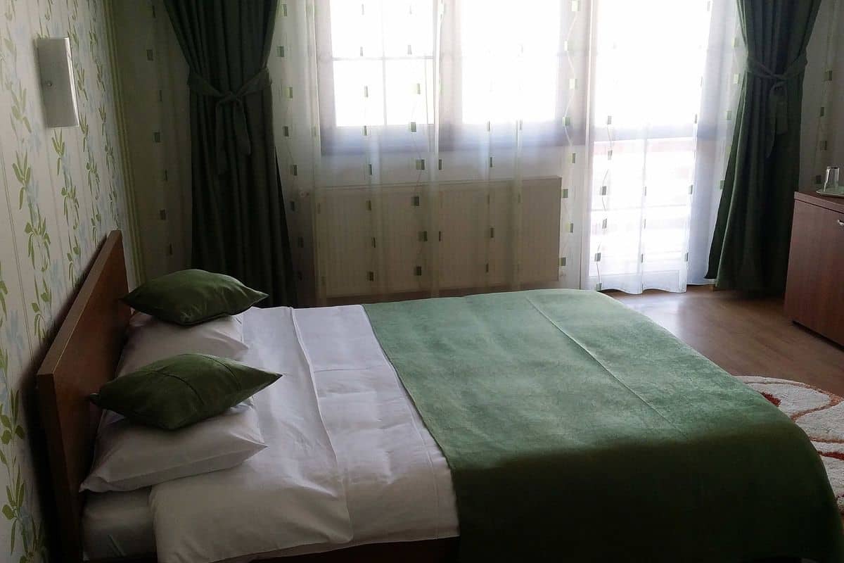 A neatly made bed with a green blanket and pillows, a wooden headboard, and a window with curtains.