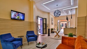 Un hall moderno de hotel con televisión, sillones azules, un sofá naranja y una zona de comedor.