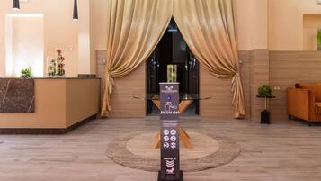 A hotel reception area with a reception desk, a sign for 'Alcohol Gel', and a 'Merci de patienter' sign on the floor.