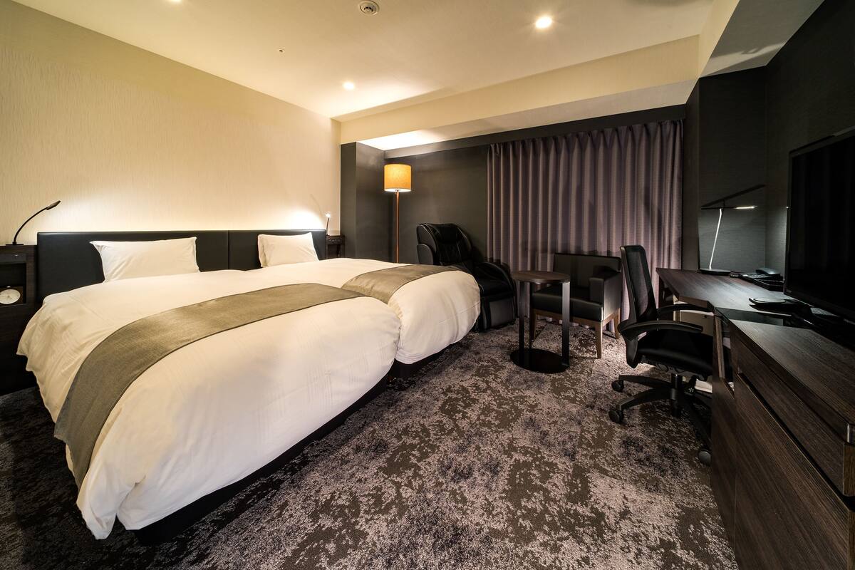 Habitación de hotel con dos camas, un escritorio, una silla, un televisor y un sillón de masaje.