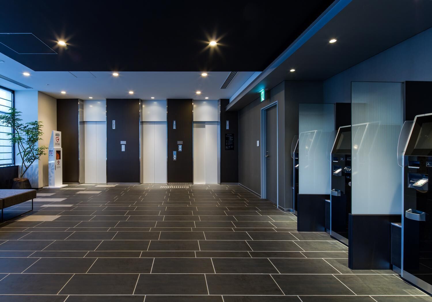 A modern hotel lobby with a tiled floor, an AED emergency station, and a reception area with a digital kiosk.