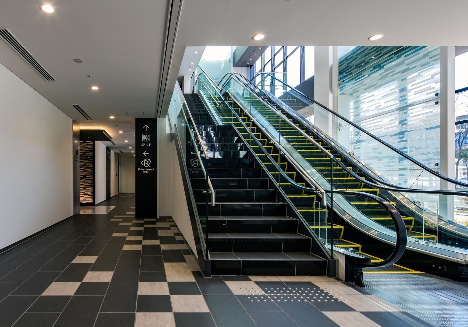 Un moderno vestíbulo de hotel con piso a cuadros, escalera de cristal y señalización direccional.